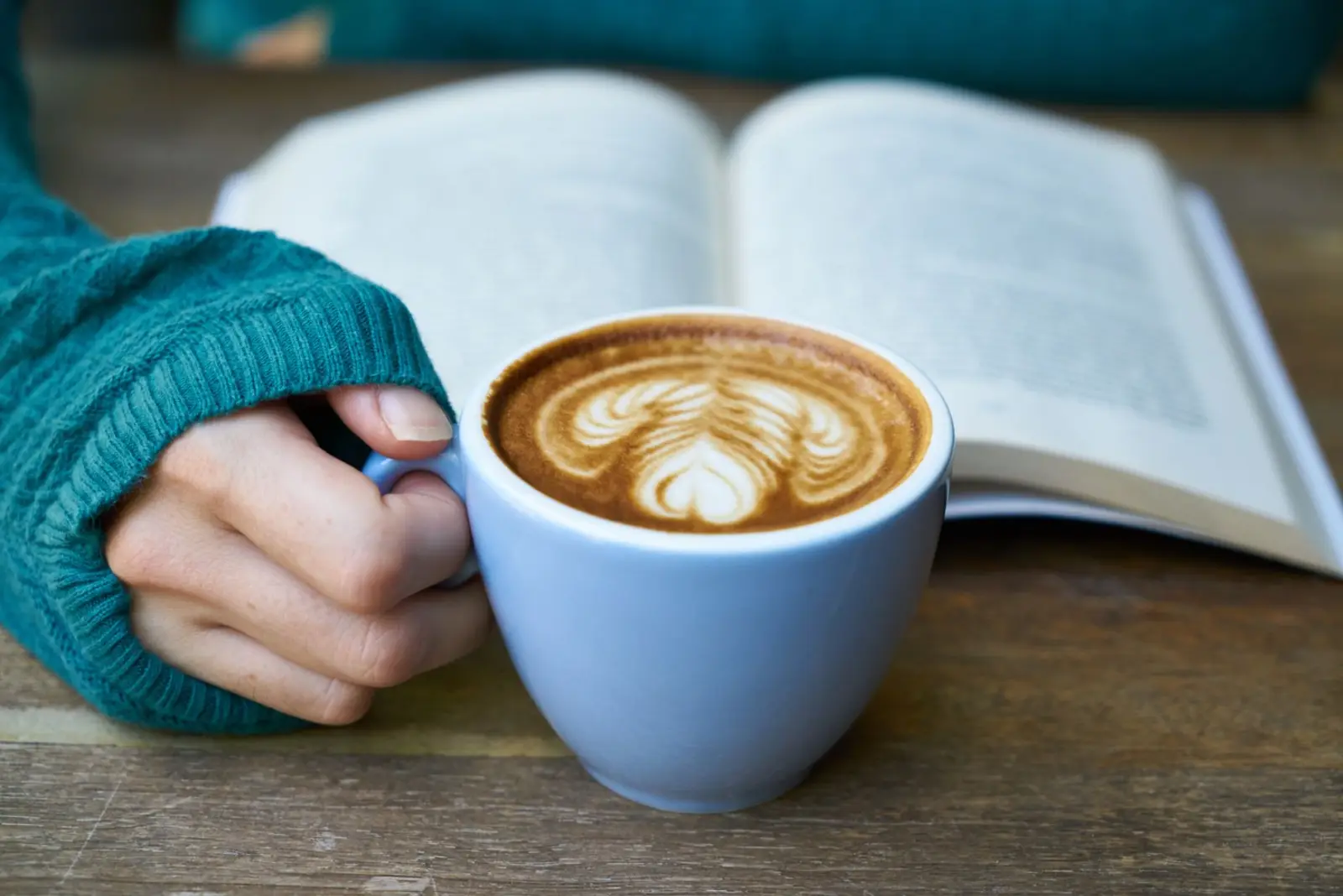 A hand holding a cappuccino next to an open book on a wooden table
